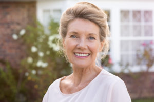 A photograph of a happy woman in her 60's smiling at the camera while standing in front of her house.