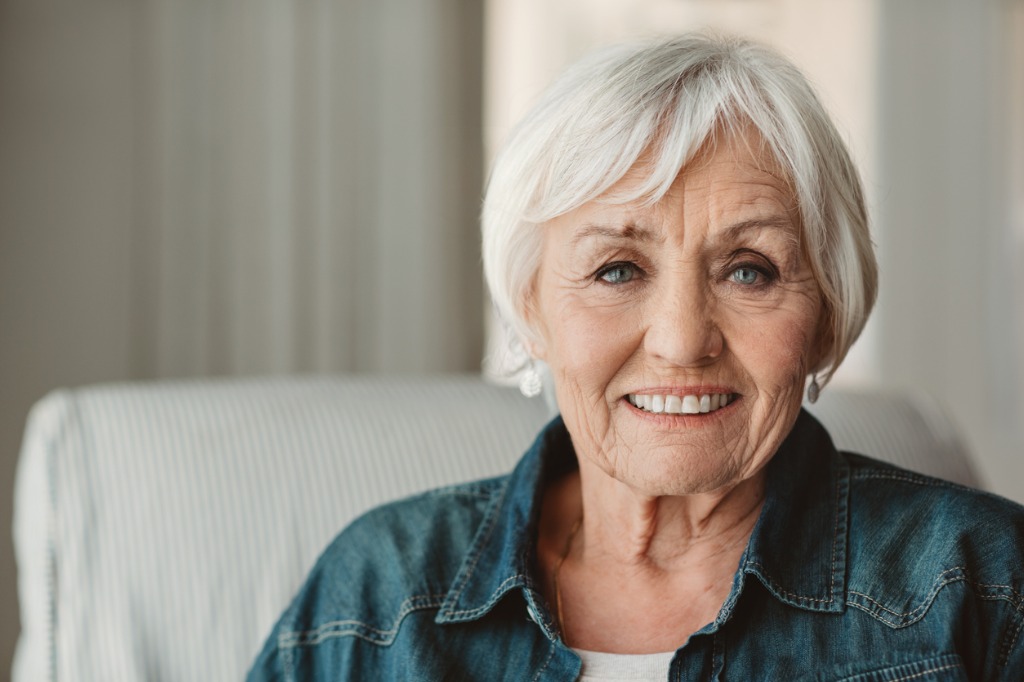 Portrait of a smiling senior woman sitting on a chair in her living room at home. She has saggy eyebrows, a prime candidate for blepharoplasty.